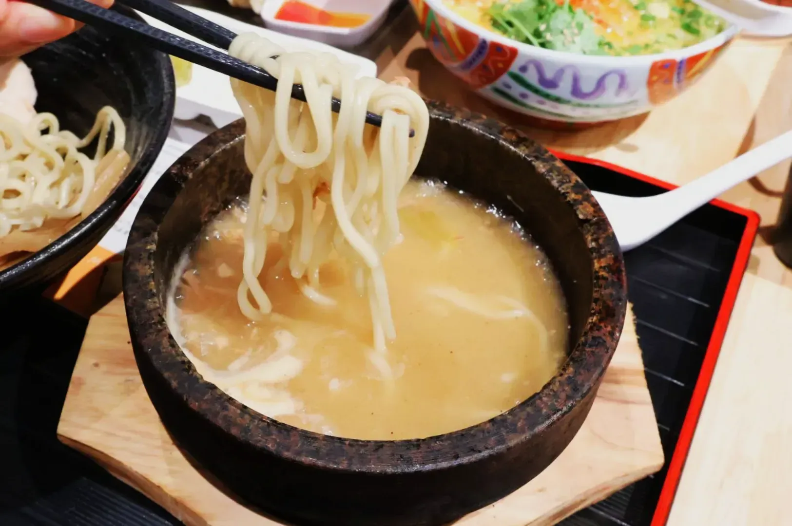 Chopsticks lift noodles from a bowl of creamy broth in a rustic stone bowl. A colorful salad and white spoon are visible in the background, creating a cozy dining scene.