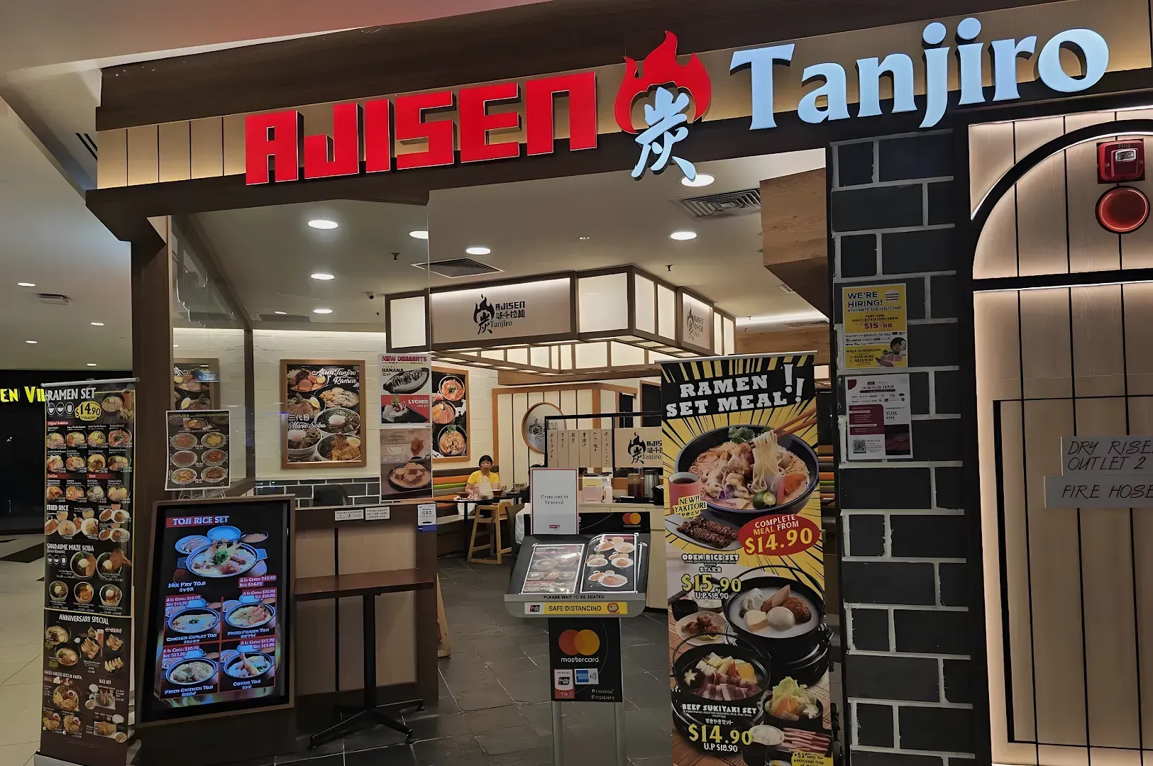 Entrance of Ajisen Tanjiri restaurant with bright signage. Menus displaying ramen set meals are visible. Cozy interior and seated diners can be seen.