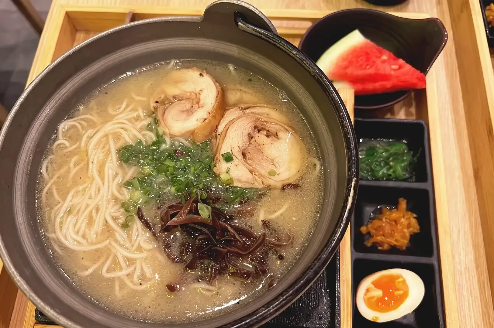 Ramen noodles in light colored broth topped with chashu, black fungus and green leaves with extra toppings and watermelon on the right side