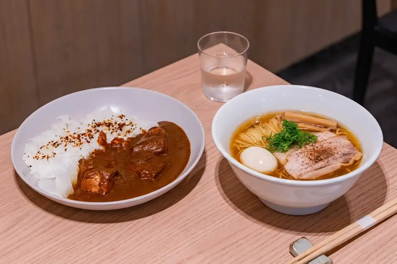 Beef curry rice with chili flakes next to a bowl of ramen with egg and bamboo shoots on a light wooden table