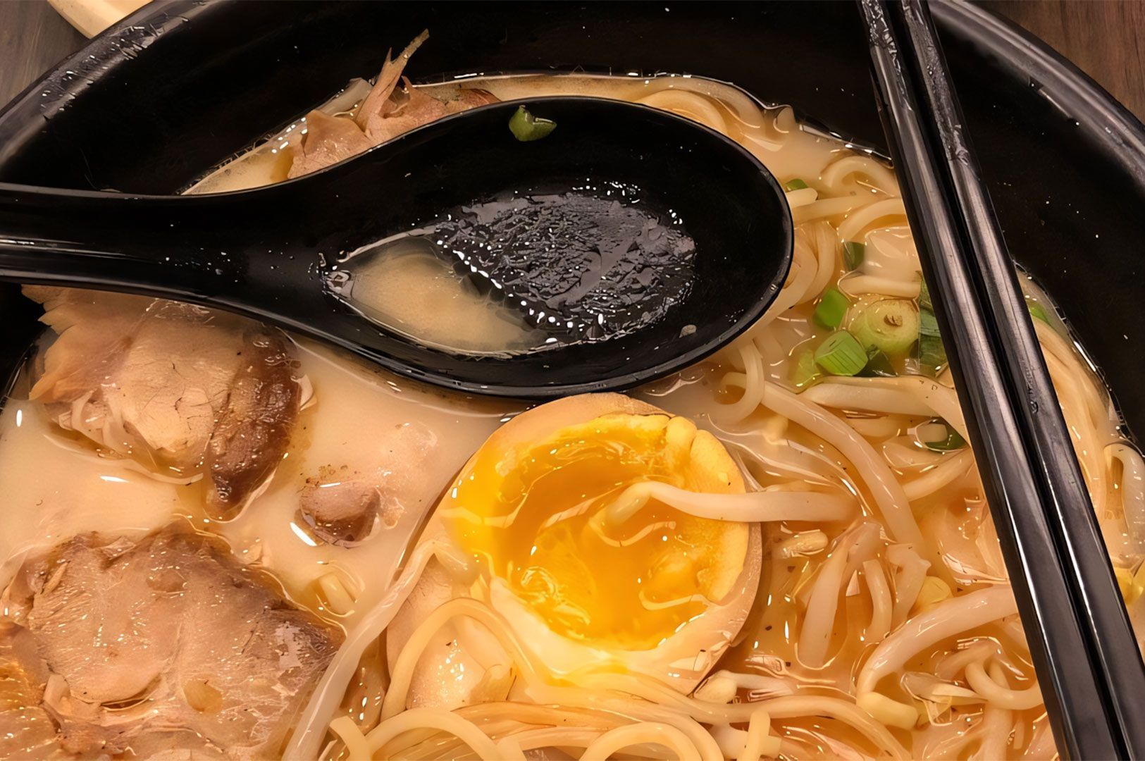Extreme close-up of a rich Tonkotsu ramen bowl with creamy broth, thick noodles, chashu pork, bean sprouts, and a soft-boiled egg with a bright orange yolk, alongside a black soup spoon.