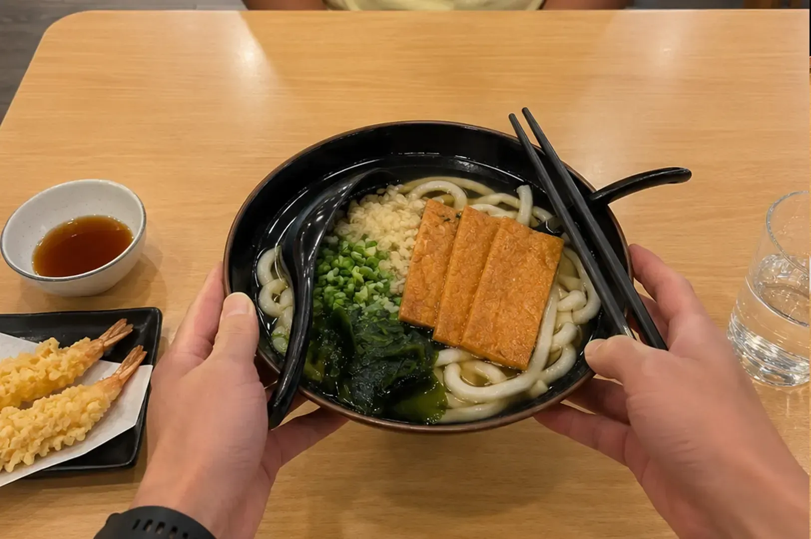 Hands holding a black bowl of udon noodles topped with fried tofu strips, green onions, and seaweed, with a side of shrimp tempura visible on the table