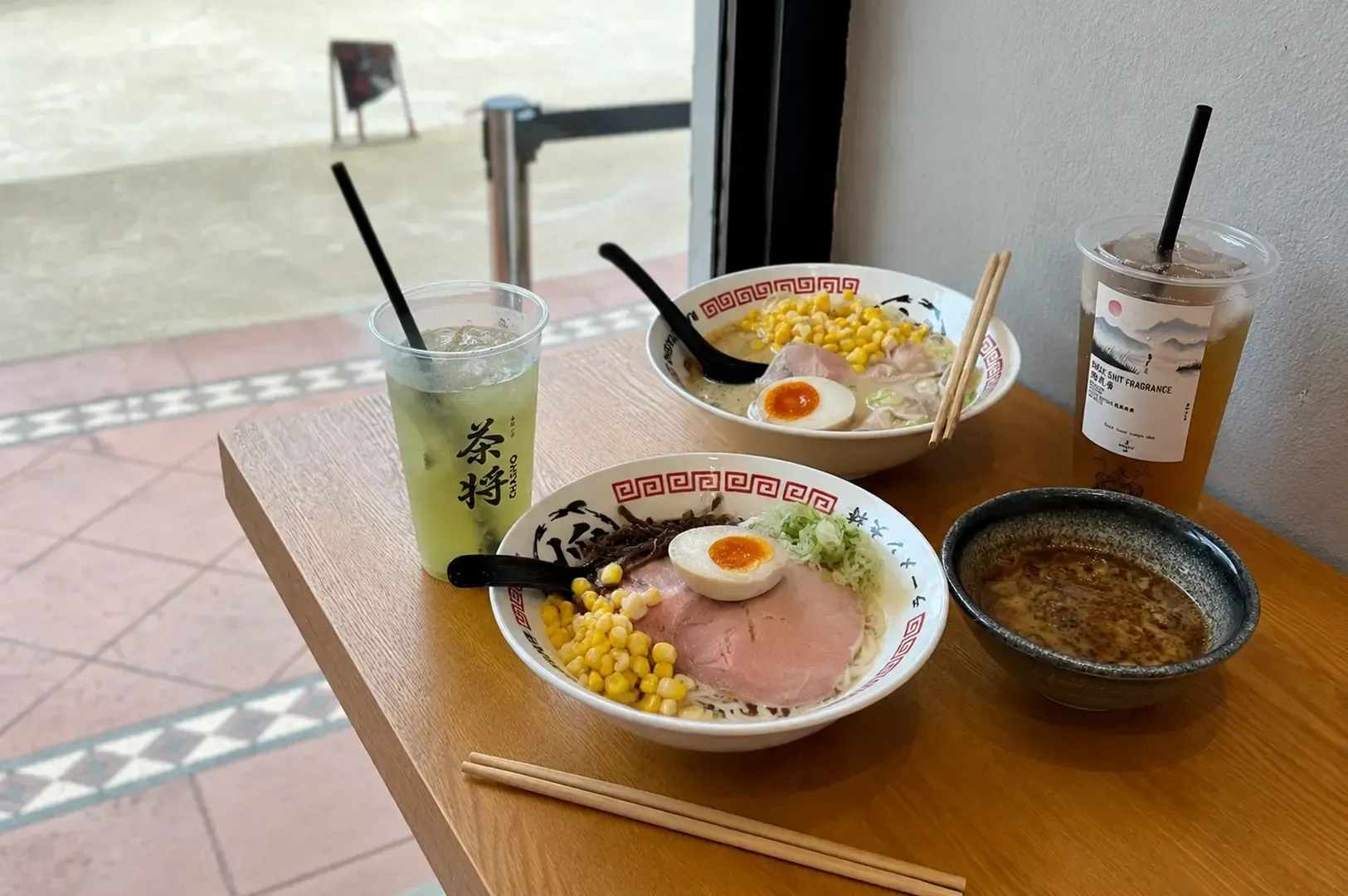 Wooden table setting at Ramen Taisho with two bowls of ramen topped with corn and egg, iced tea drinks, and a small side bowl near a window