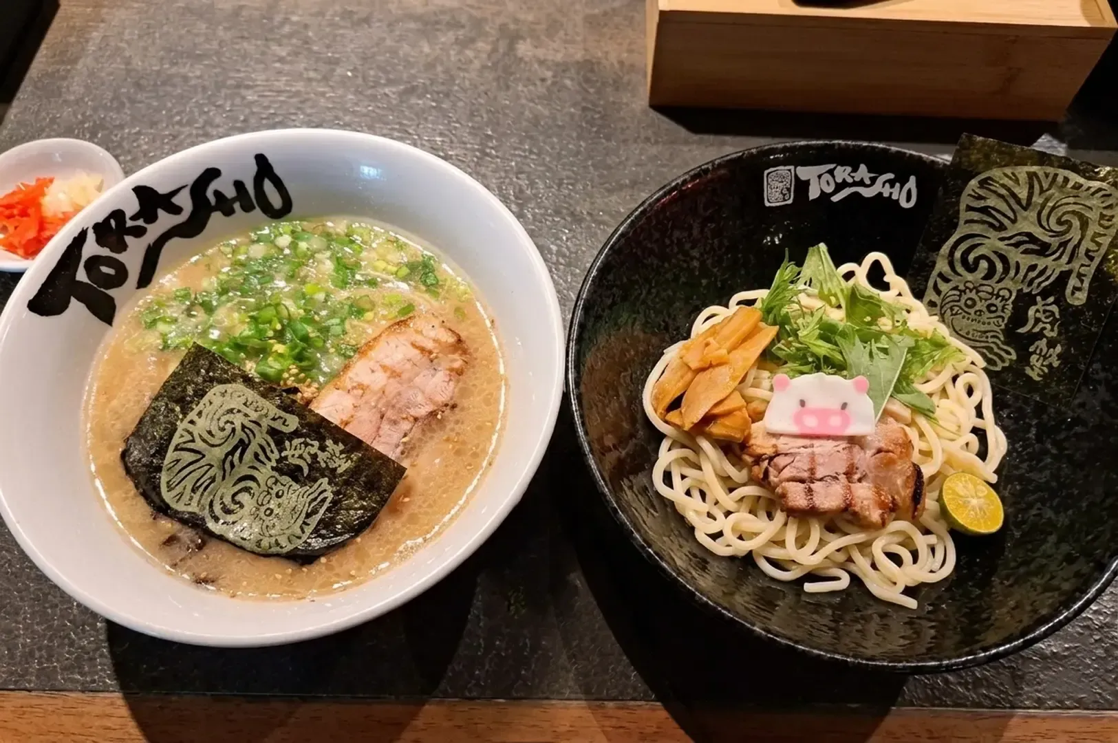 Two Torasho ramen bowls side-by-side, one with creamy broth and the other a dry noodle dish featuring a pig-shaped fish cake and tiger-branded nori