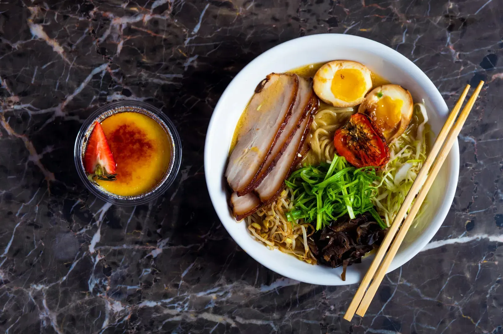 A white bowl of ramen with sliced pork, halved eggs, green onions, and seaweed, on a marbled surface, accompanied by crème brûlée with a strawberry slice.
