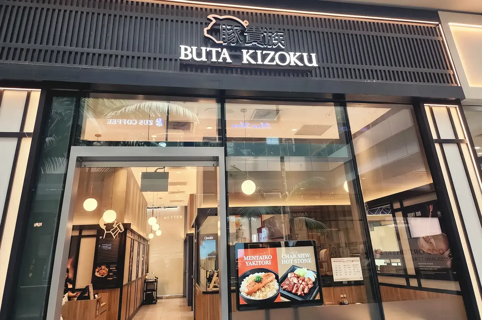 Sleek black and beige themed storefront of Buta Kizoku peeking through warm lighted interior and food posters