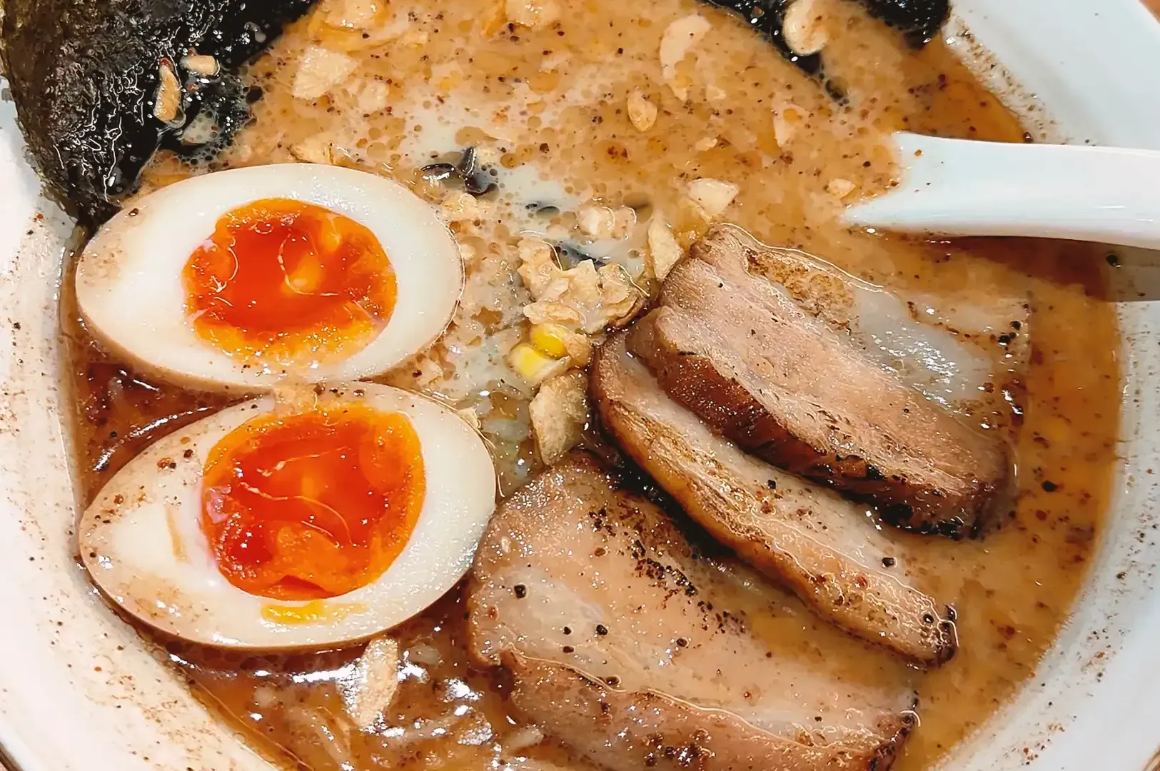 Ramen bowl containing slices of roasted pork, eggs in half, corn, garlic flakes and seaweed submerged in broth mixed with black sauce