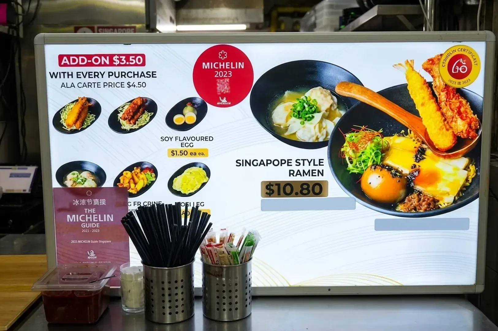 Illuminated menu board for A Noodle Story displaying Singapore Style Ramen for $10.80 and Michelin 2023 recognition.