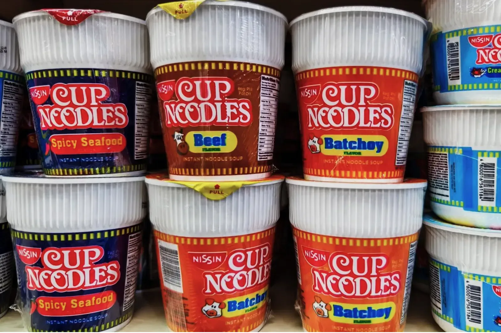 Close-up of Nissin Cup Noodles on a store shelf. Visible flavors: Spicy Seafood, Beef, Batchoy. Bright packaging with bold text creates a vibrant display.