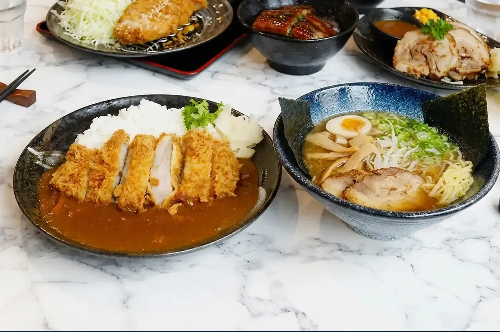 hicken katsu curry rice and a bowl of ramen with egg and nori on a white marble table surrounded by other Japanese dishes