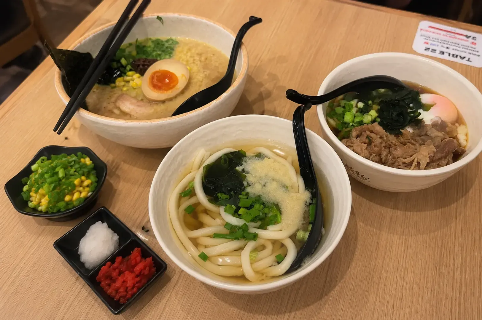 Table spread featuring bowls of creamy ramen, beef udon, and clear broth udon accompanied by small condiment dishes