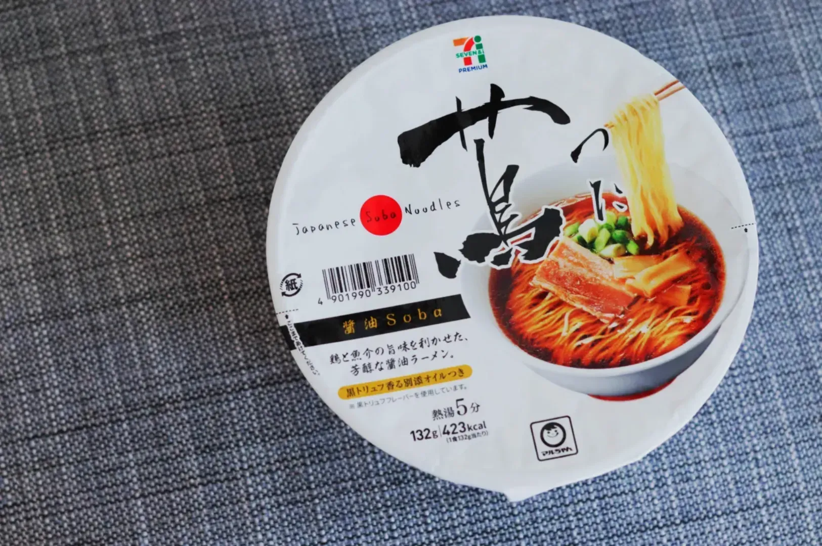 A top view of packaged Japanese soba noodles on a gray surface. The packaging features bold calligraphy, an image of noodles in broth, and the 7-Eleven logo.
