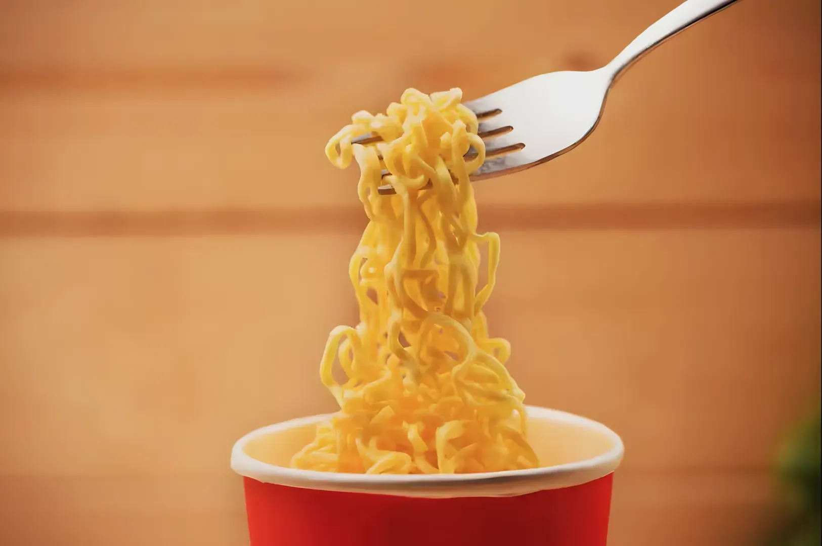 A fork lifts steaming yellow noodles from a red cup against a blurred wooden background, conveying warmth and simplicity. Keywords: noodles, fork, cup.