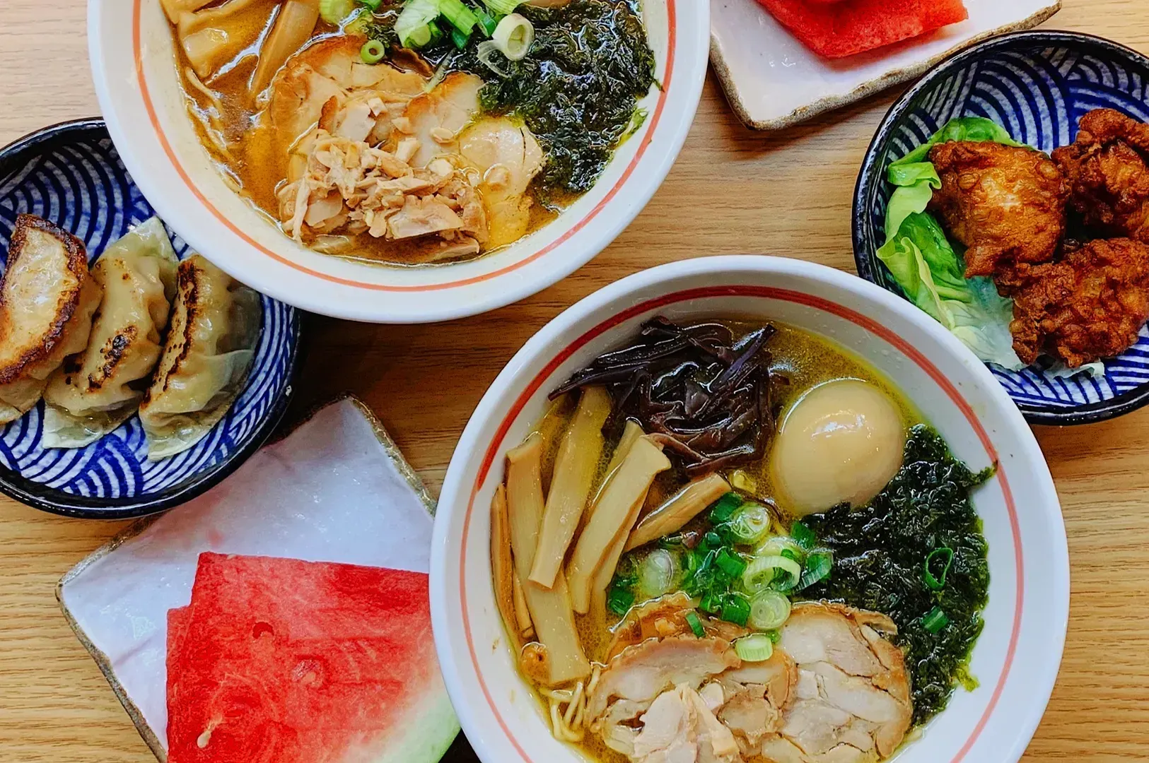 Two bowls of ramen with sliced meat, seaweed, scallions, and eggs. Sides include gyoza, fried chicken, and watermelon, arranged on a wooden table.