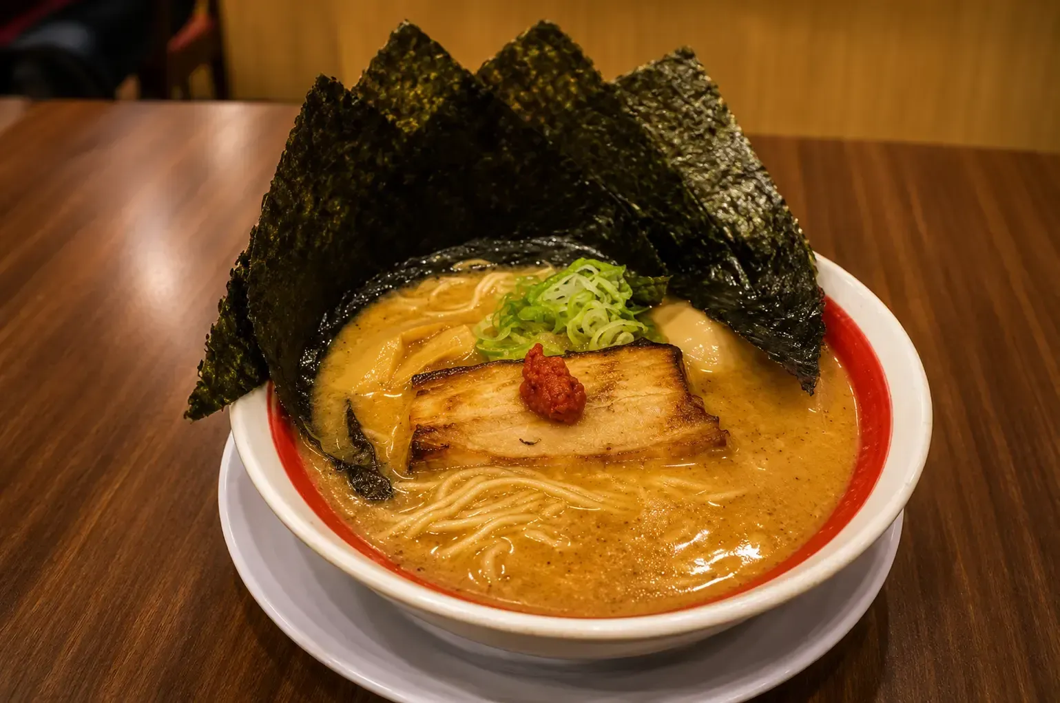 Bowl of ramen in rich broth topped with a thick slice of grilled pork belly, green onions, and large sheets of nori seaweed.