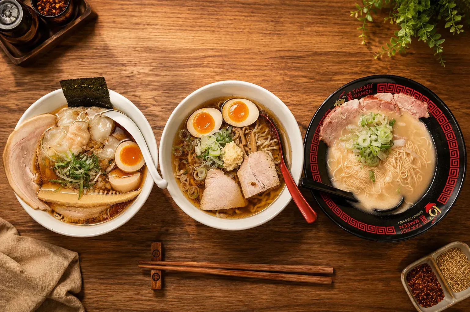 A chef in a black uniform creates a massive burst of flame while pouring hot oil from a pan into a bowl of ramen on a wooden counter, as customers watch from the left in a Japanese ramen restaurant.