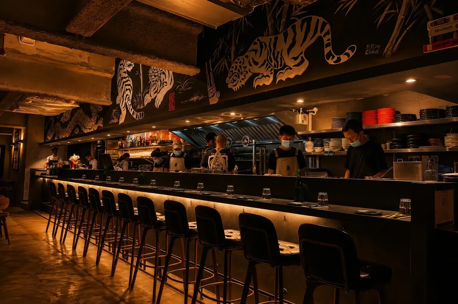 A dimly lit, modern restaurant featuring a long bar counter with sleek leather barstools. Chefs in masks work in an open kitchen behind the counter, under a dark soffit decorated with large white paintings of tigers and dragons.