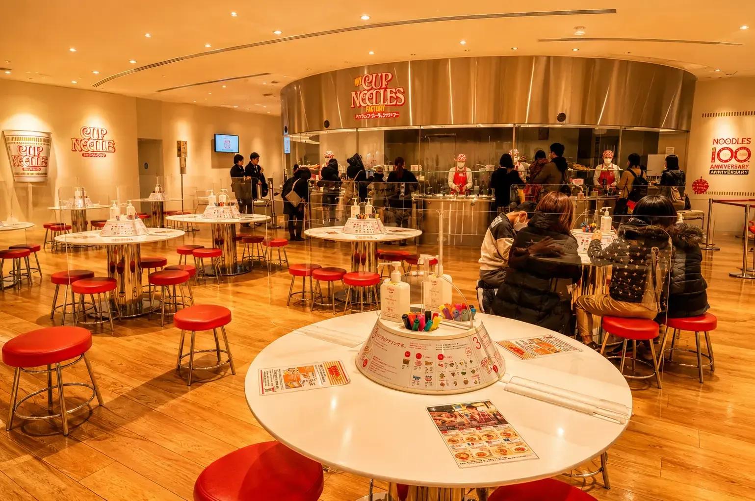 An indoor workshop space at the My Cup Noodles Factory, featuring round tables with red stools where people are decorating their own noodle cups, with a glass-enclosed kitchen counter and staff in the background.