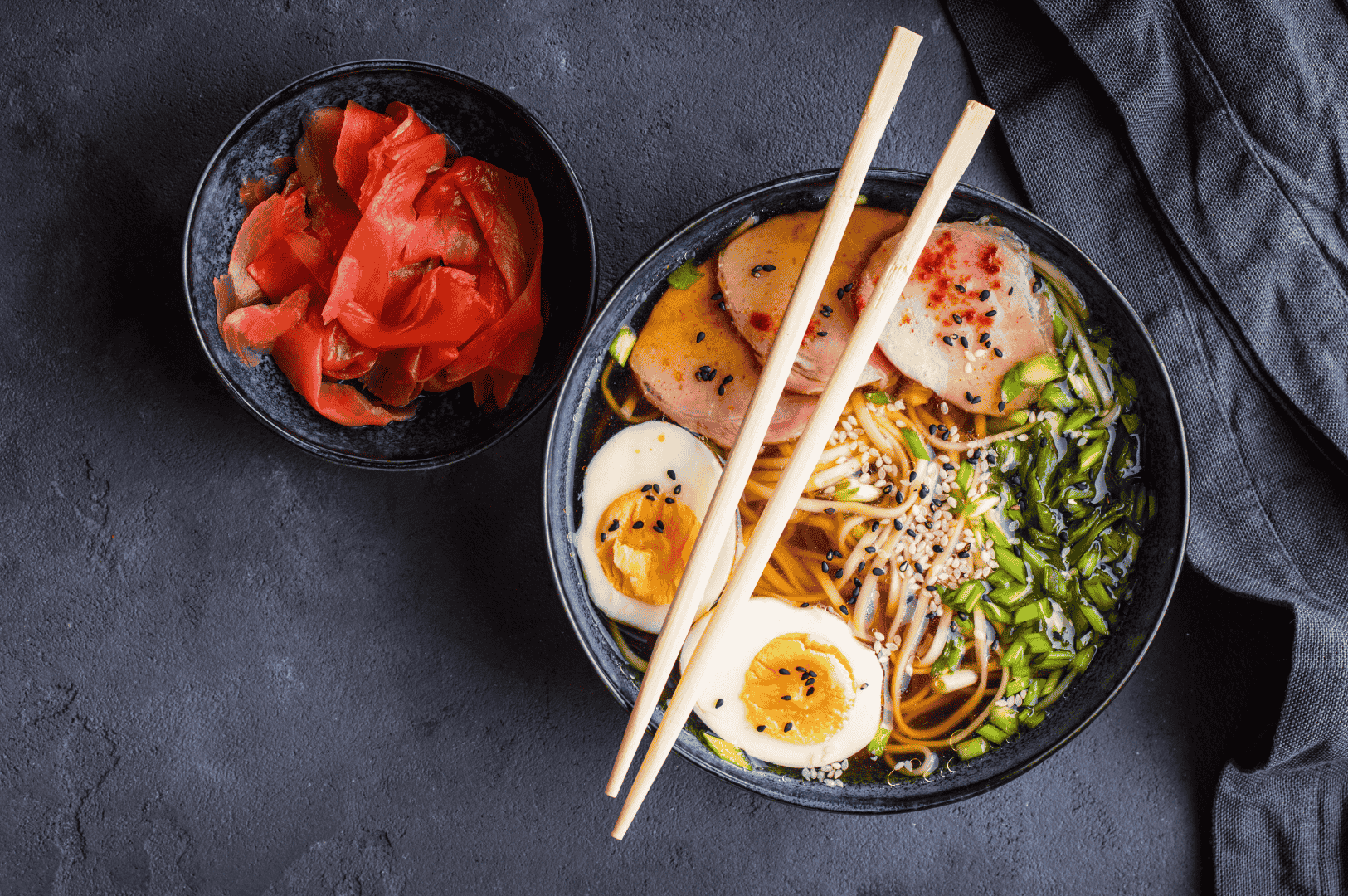 Top-down view of a garlic-forward ramen bowl featuring rich broth, tender pork slices, jammy eggs, scallions, sesame seeds, and springy noodles, with crispy garlic notes subtly layered throughout. A side of pickled ginger adds contrast, while wooden chopsticks and a dark napkin frame the composition—capturing the depth and nuance of garlic infusions in Japanese comfort cuisine.