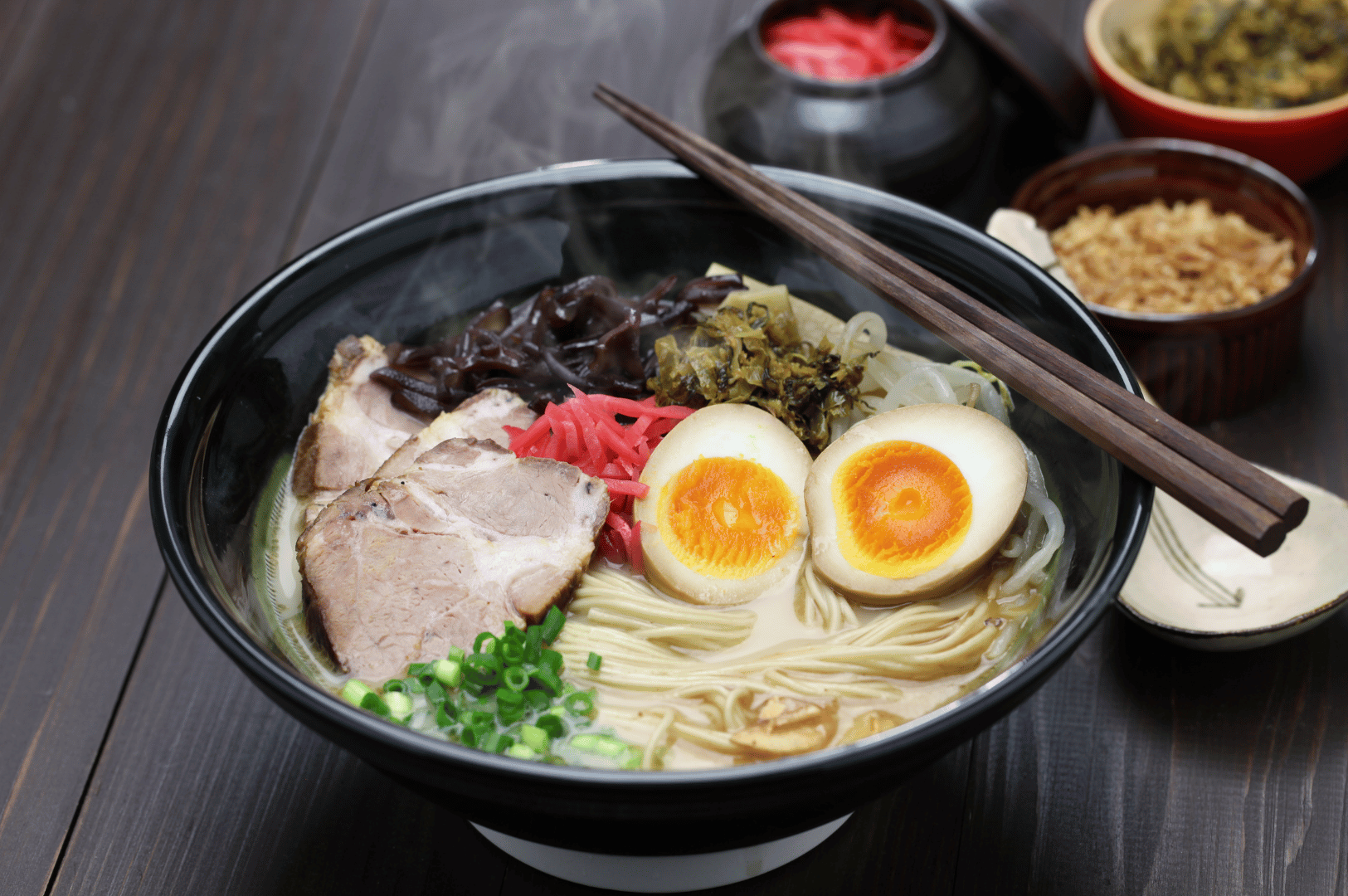 Steaming bowl of ramen in a black vessel, layered with chashu pork, jammy eggs, wood ear mushrooms, pickled ginger, mustard greens, bean sprouts, and scallions—all immersed in a creamy garlic-infused broth. Chopsticks rest across the bowl, while background condiments like crispy garlic and pickled ginger hint at customizable depth, capturing the sensory richness and cultural artistry of garlic-forward ramen.