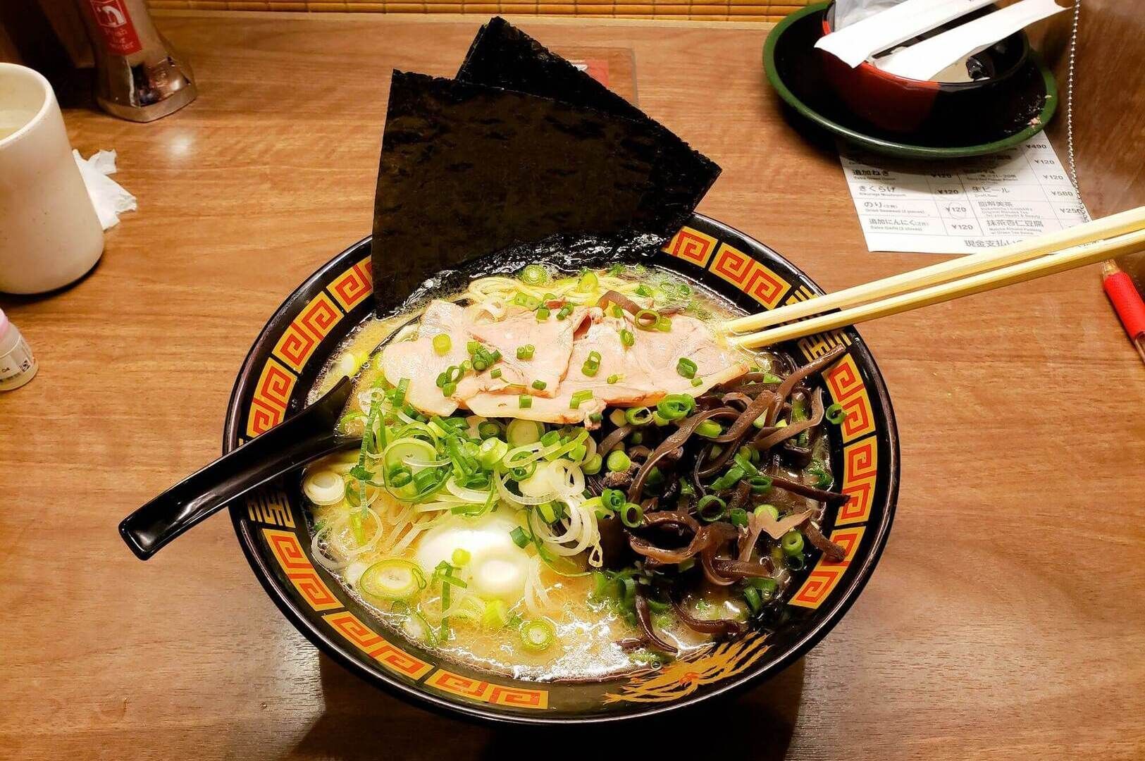 A black plate holds a bowl of Hakata ramen, accompanied by chopsticks, highlighting the dish's savory ingredients.