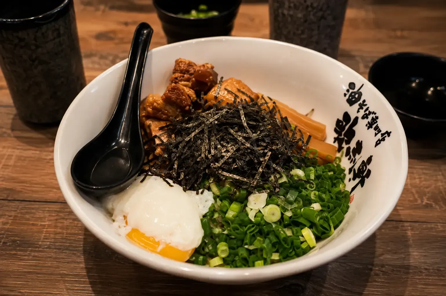 A bowl of dry ramen topped with braised pork belly, poached egg, bamboo shoots, shredded seaweed, and chopped green onions.