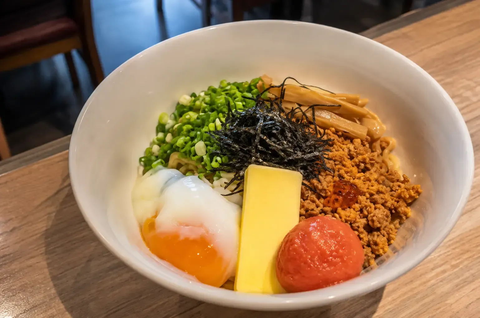 Close-up of a bowl of dry ramen topped with minced pork, soft-boiled egg, butter, spicy cod roe, bamboo shoots, and green onions.