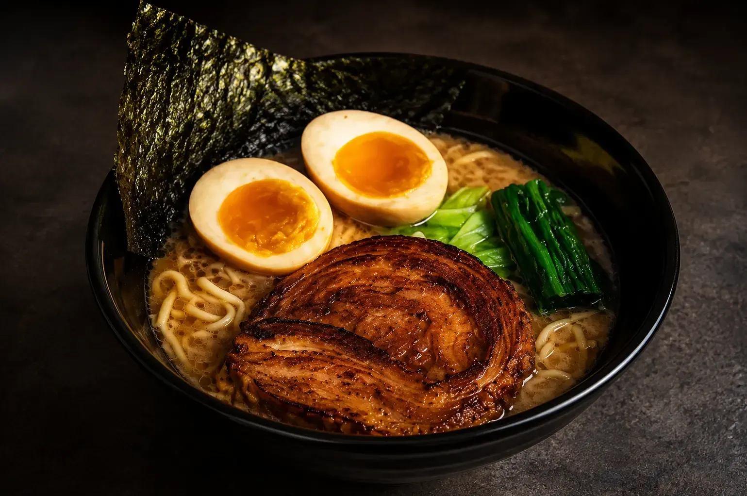 A bowl of ramen featuring a large slice of rolled seared chashu pork, soft-boiled eggs, greens, and a crisp sheet of nori.