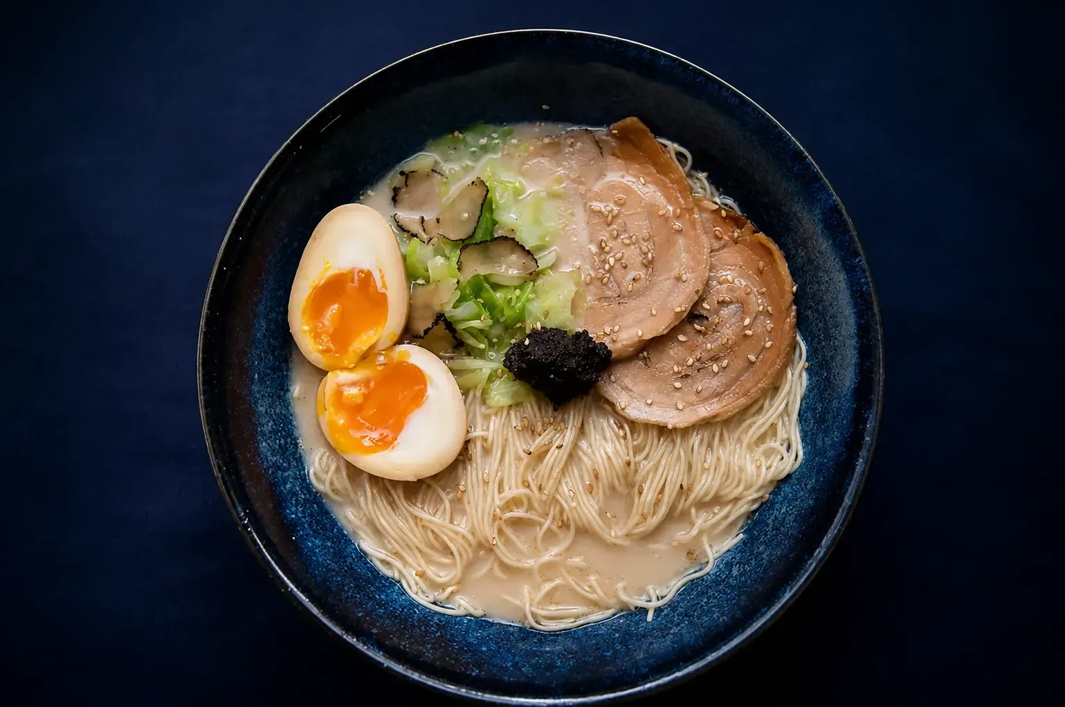 Top-down view of a bowl of truffle ramen topped with thin noodles, sliced chashu pork, soft-boiled eggs, shaved truffle, and black truffle paste.