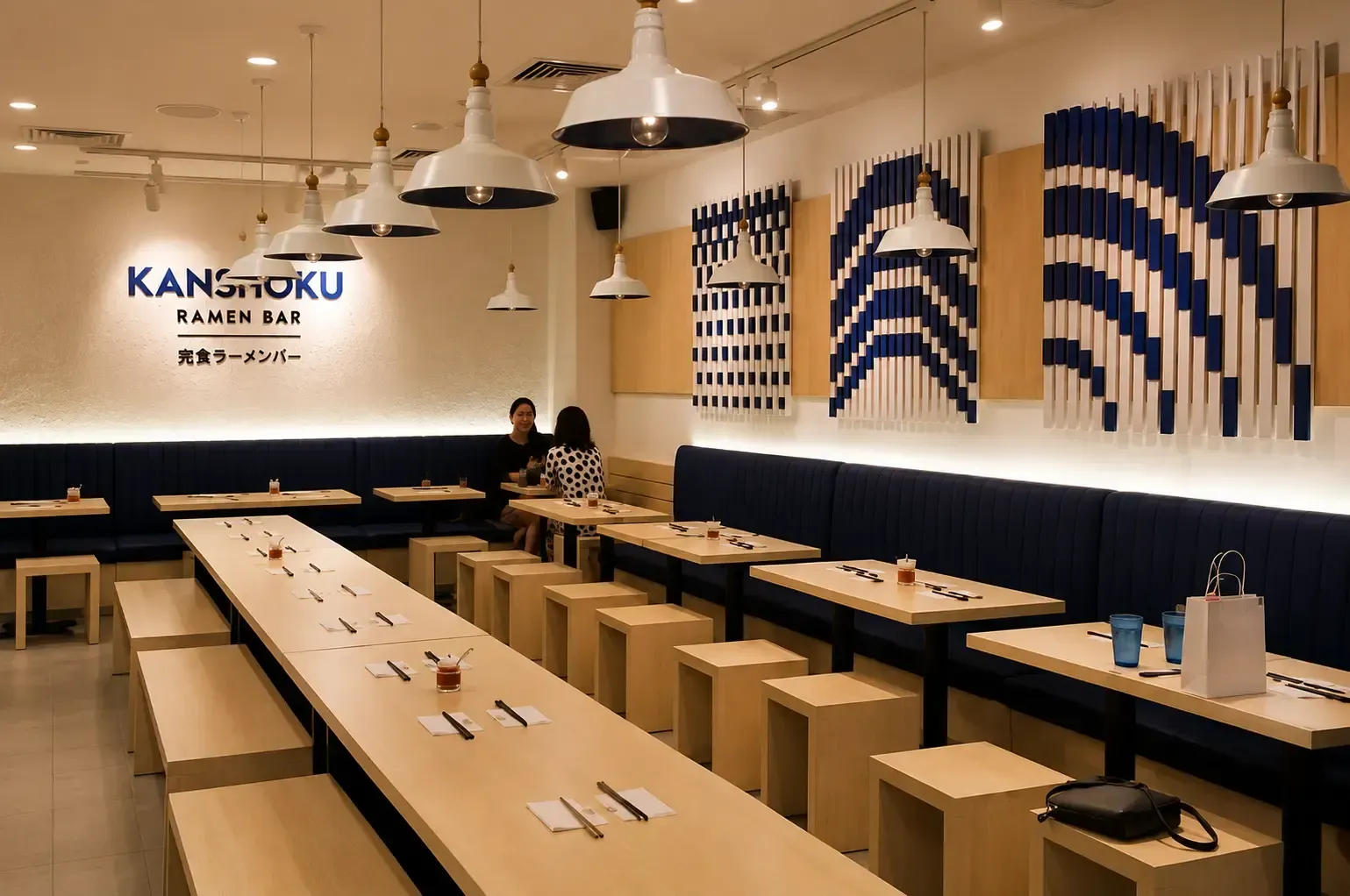 Clean interior view of Kanshoku Ramen Bar showing long light-wood communal tables, stool benches, and blue booth seating.