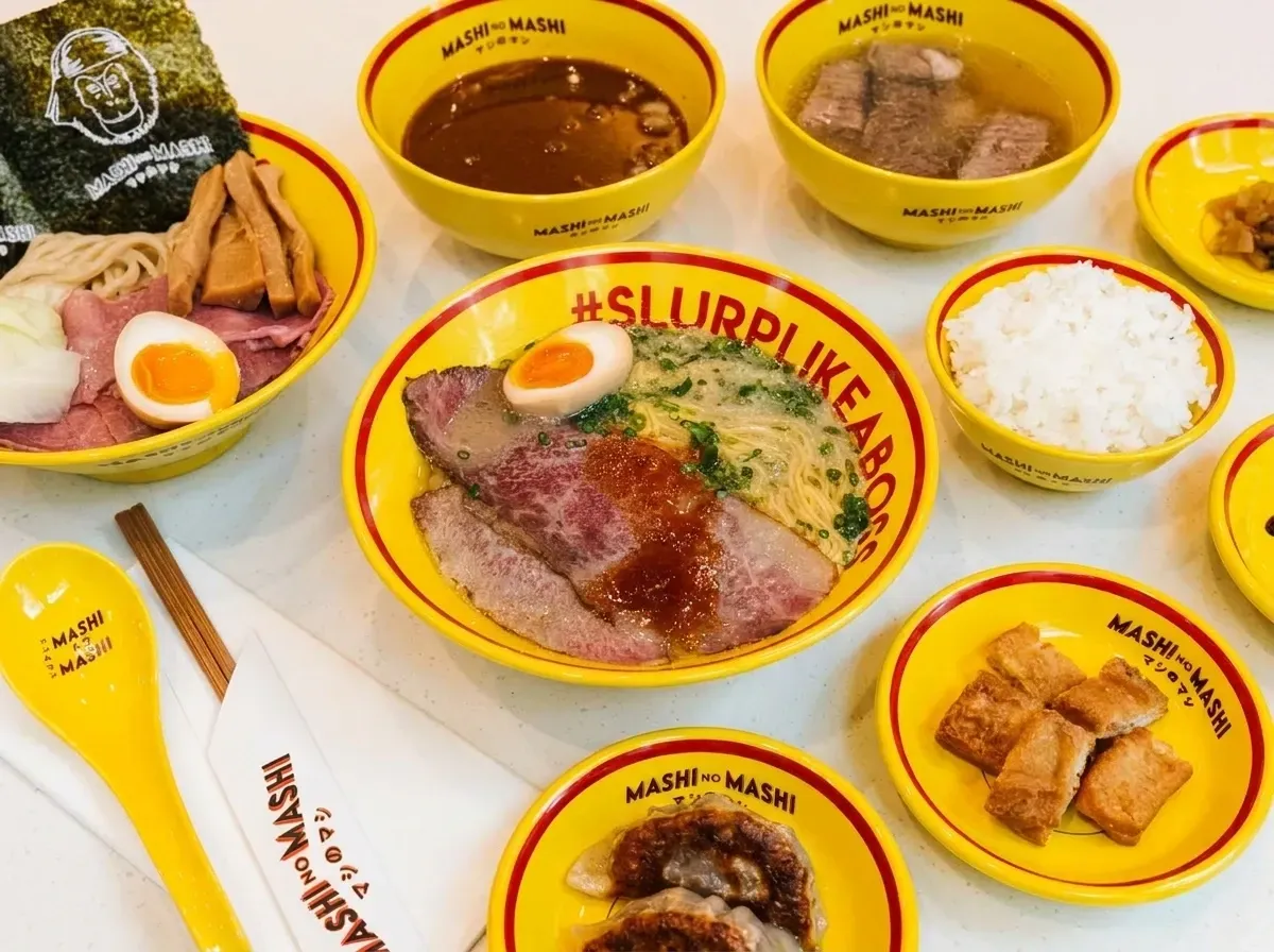 A flat lay view of a Japanese meal spread featuring a central bowl of wagyu beef ramen, surrounded by tsukemen dipping noodles, side dishes of gyoza, crispy tofu, white rice, and broth, all served in matching bright yellow branded tableware.