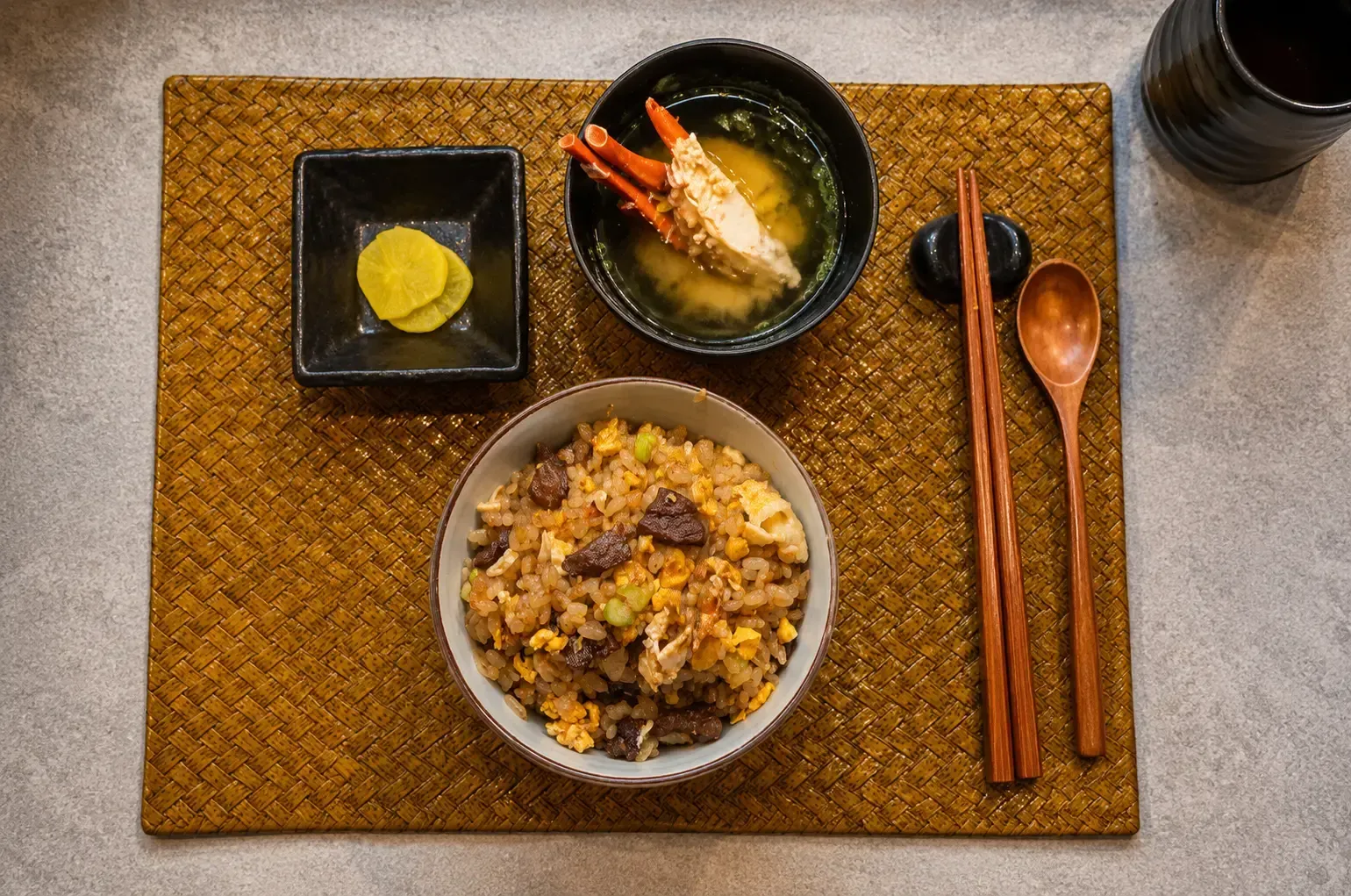 Overhead view of a fried rice bowl served with pickled radish and crab leg soup on a woven placemat.