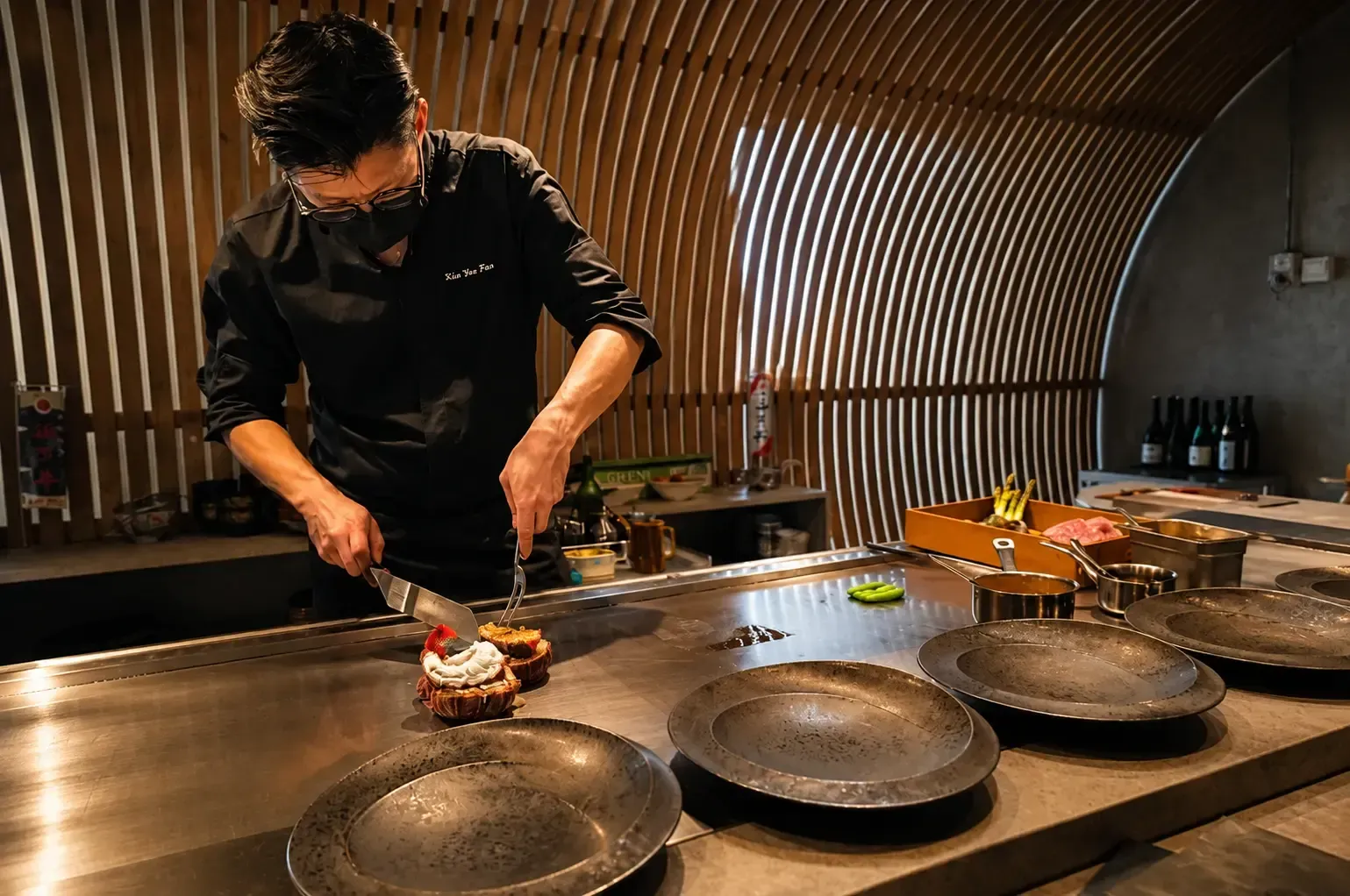 A masked chef plated dishes over a sleek stainless steel teppanyaki flat-top grill.