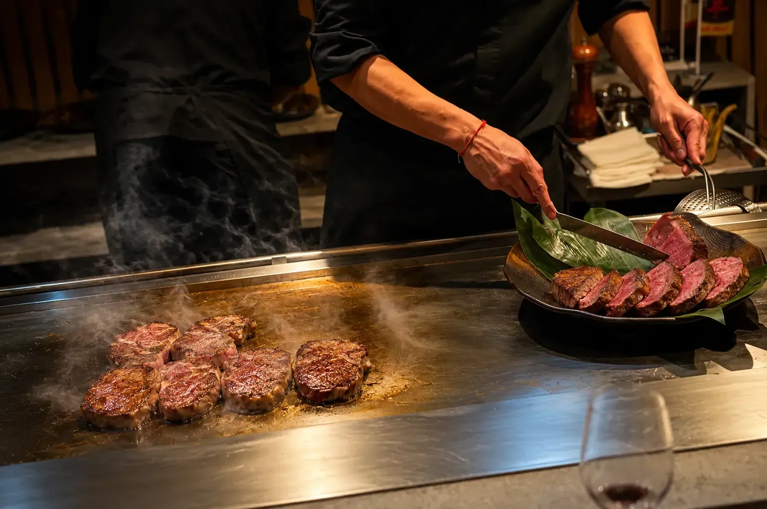 Chef searing thick-cut wagyu beef steaks on a smoking flat-top grill next to sliced cooked steak.