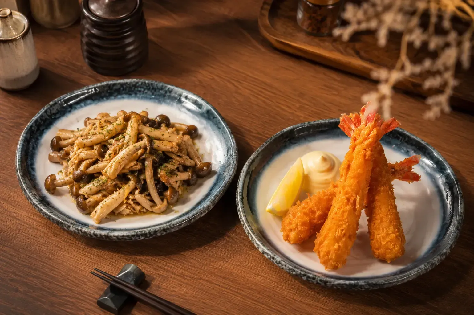 A plate of sautéed wild mushrooms and a plate of crispy fried shrimp tempura served with a lemon wedge and dipping sauce on a wooden table.
