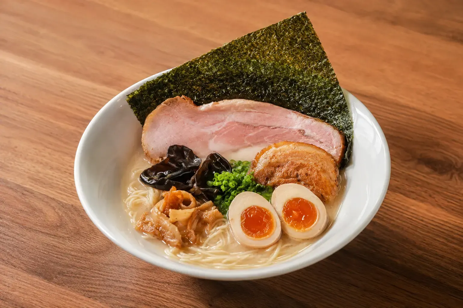 A bowl of ramen with a soft-boiled egg cut in half, sliced chashu pork, wood ear mushrooms, green onions, and a sheet of nori seaweed on a wooden table.