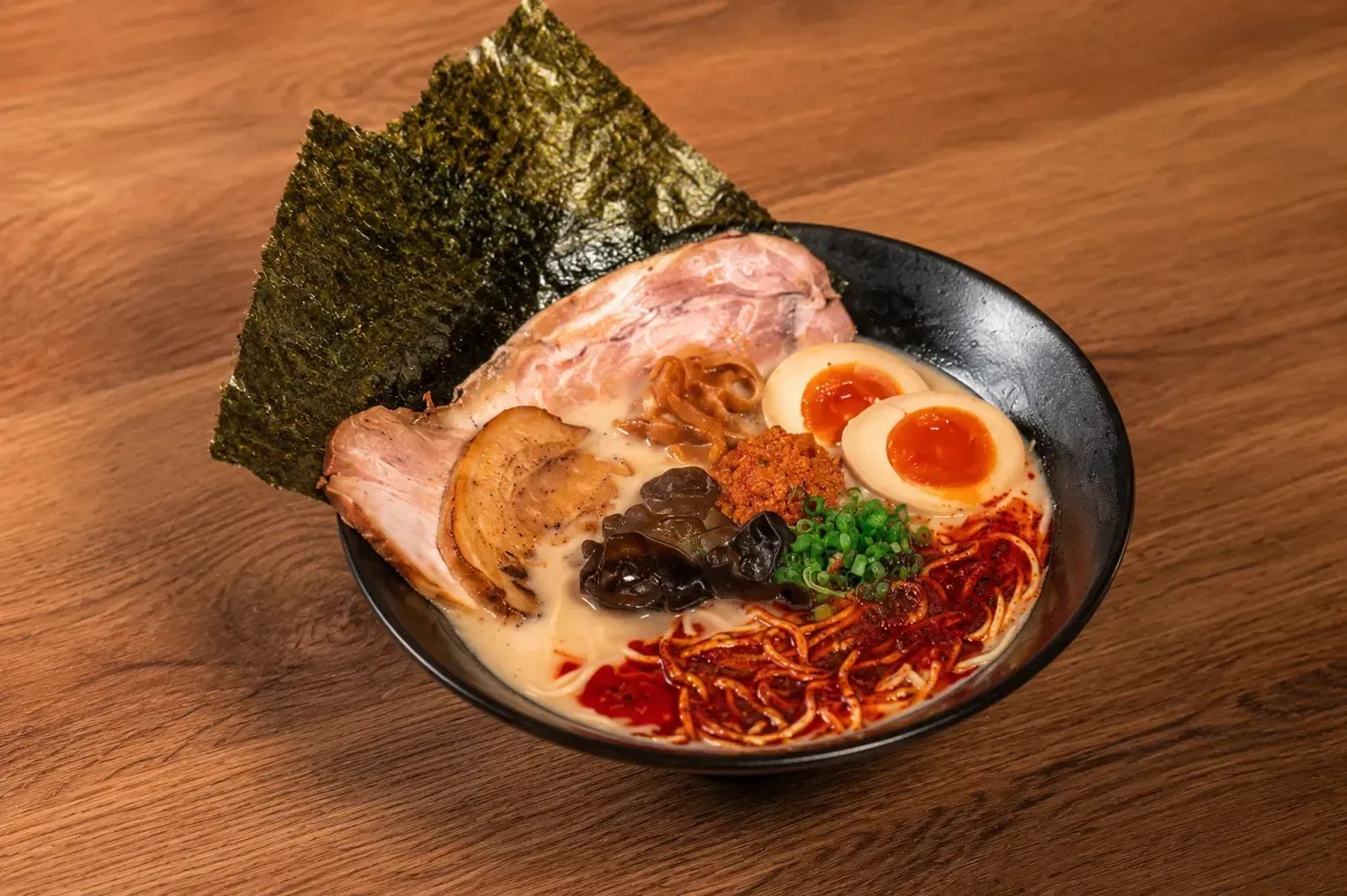 A bowl of spicy ramen in a dark bowl, topped with double chashu pork slices, soft-boiled egg halves, wood ear mushrooms, green onions, and a sheet of nori seaweed over a rich, reddish chili oil broth.