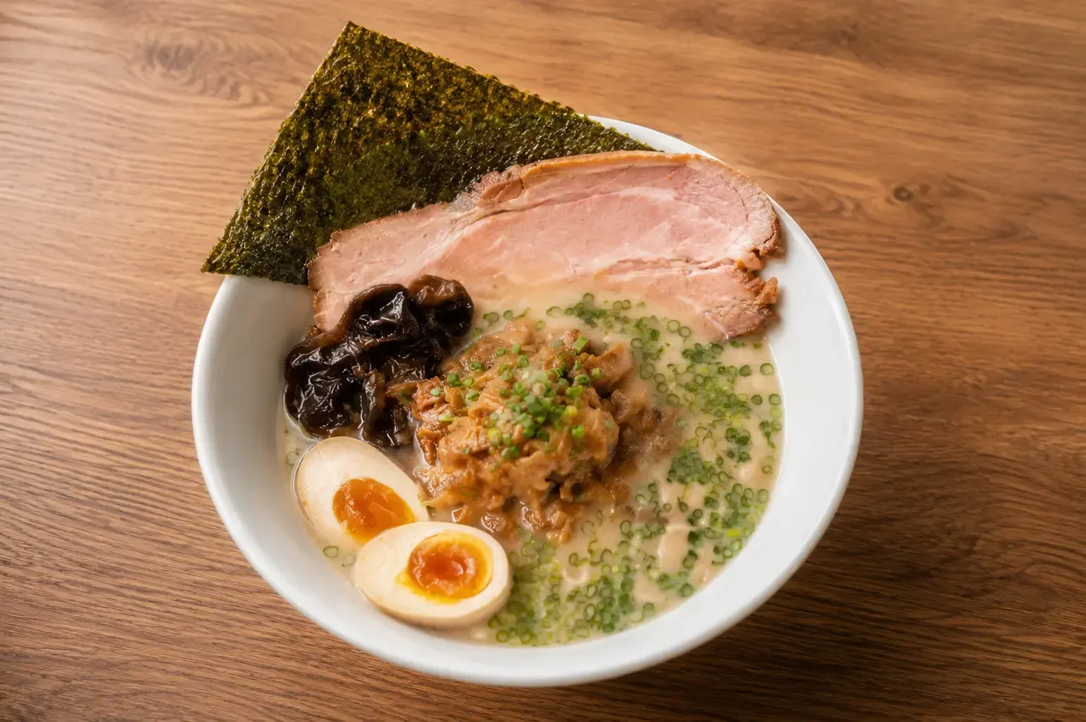 A bowl of ramen featuring tonkotsu broth, minced seasoned meat, sliced chashu pork, a soft-boiled egg, green onions, and a large sheet of seaweed.