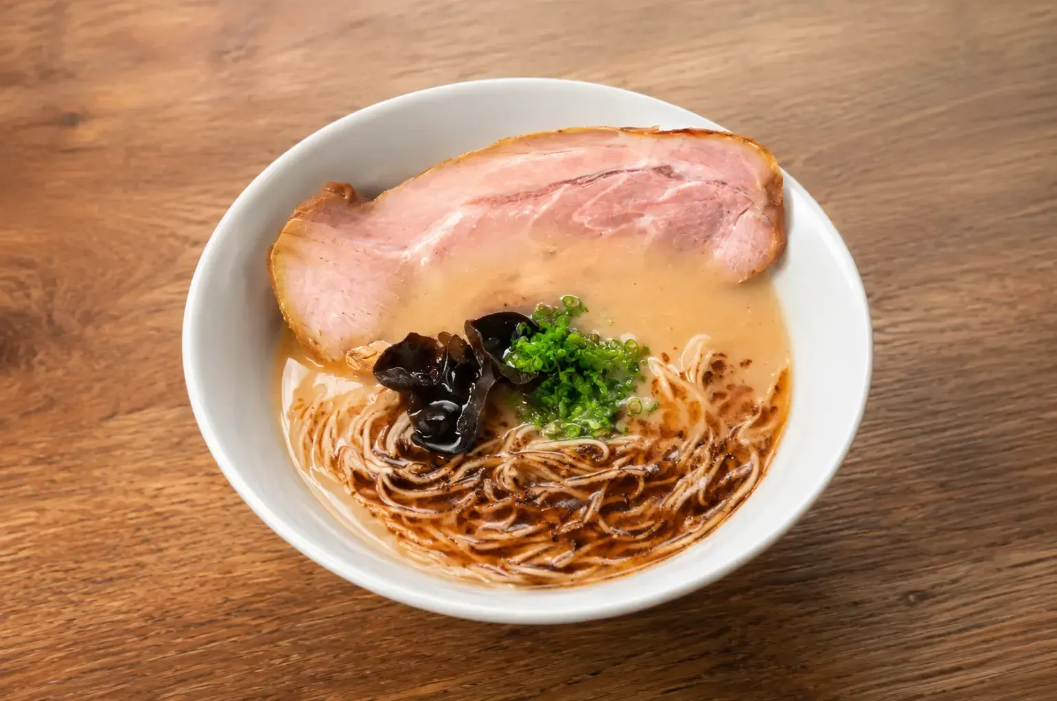 A bowl of ramen topped with a large slice of chashu pork, wood ear mushrooms, fresh green onions, and a drizzle of dark black garlic oil in a savory broth.