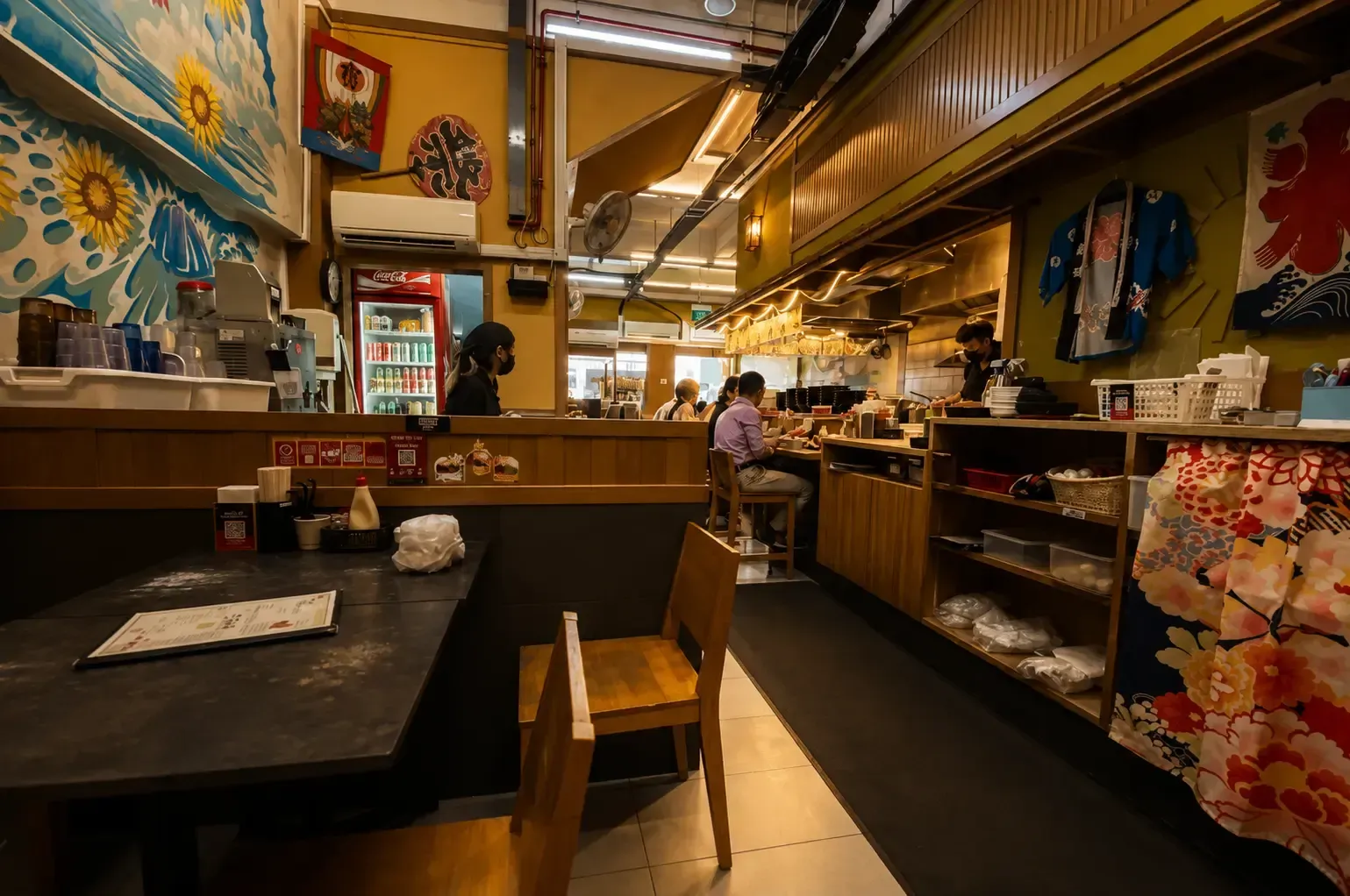 The cozy interior of a Japanese ramen restaurant featuring counter seating, colorful wall murals, and traditional decorative fabrics.