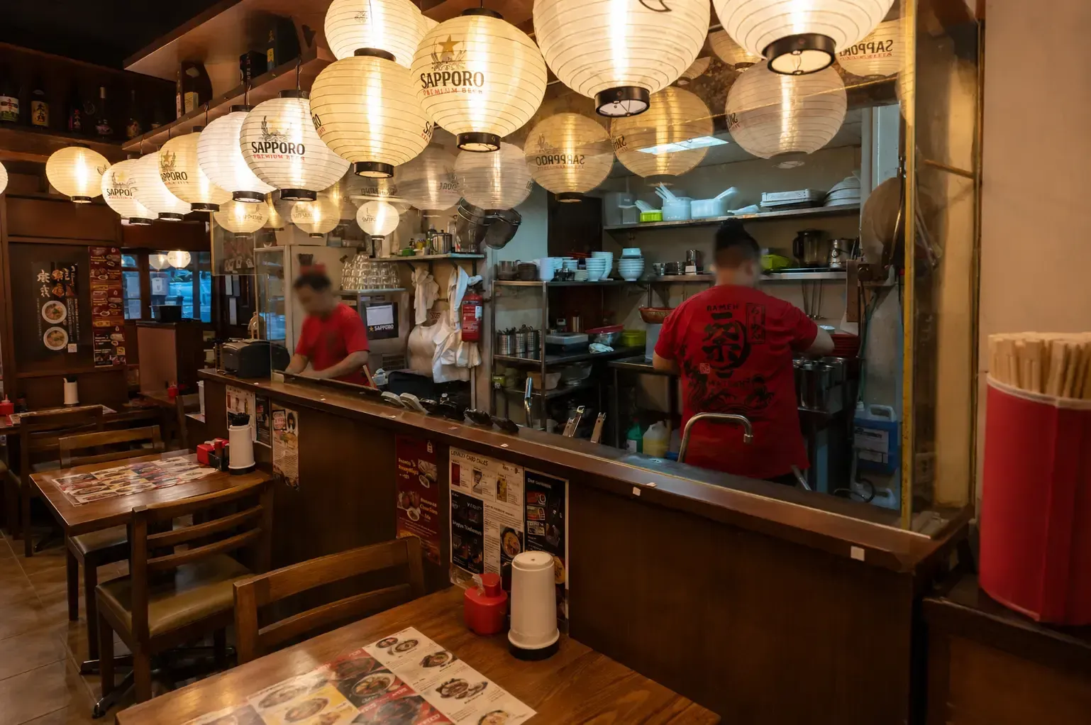 Wooden tables and an open kitchen counter inside a ramen shop under a ceiling packed with illuminated paper lanterns.