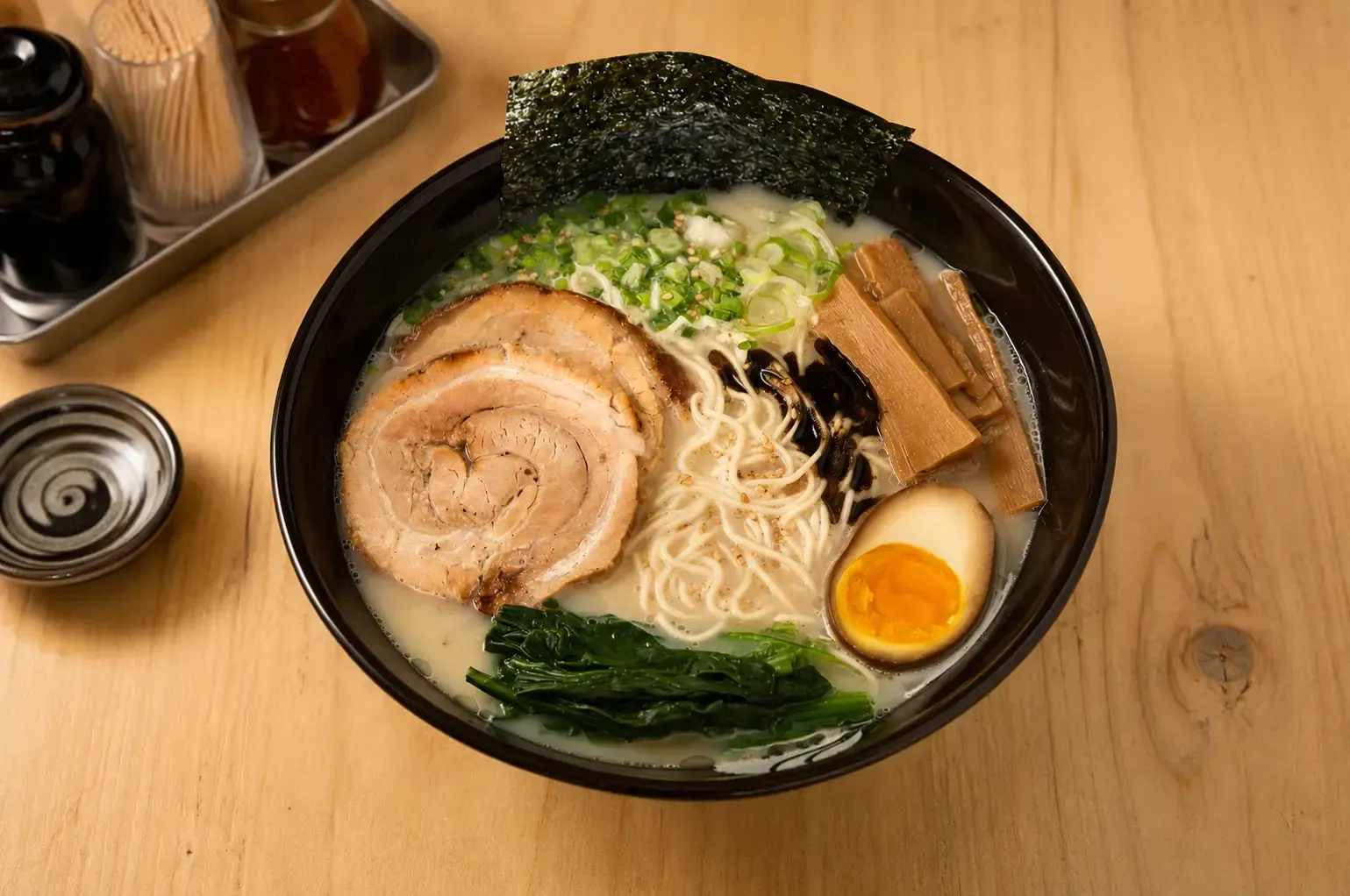 A black bowl of tonkotsu ramen with noodles, chashu pork slices, a soft-boiled egg, green onions, bamboo shoots, and nori seaweed, served on a light wooden table.