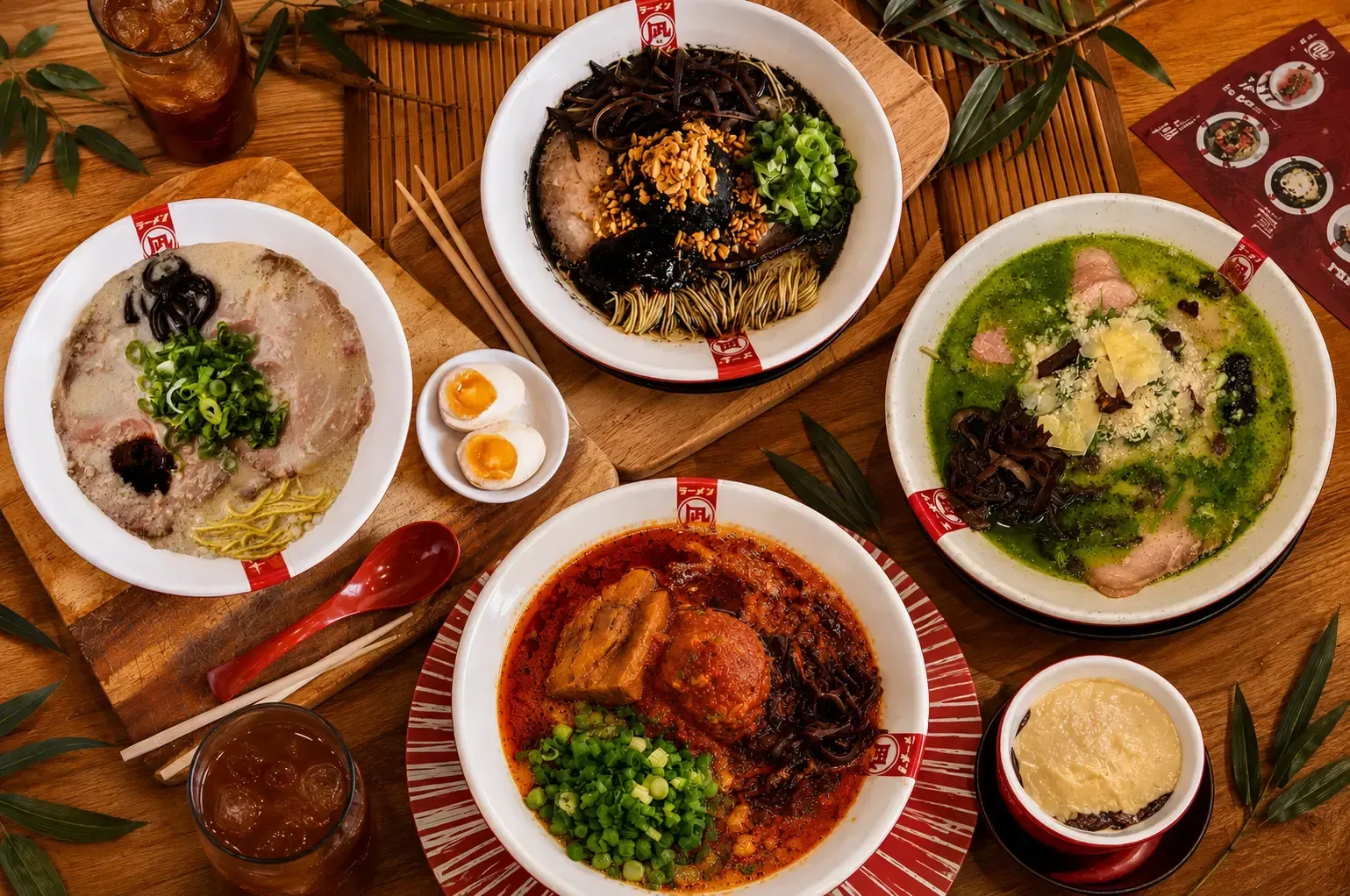 Overhead flat lay of four distinct bowls of ramen—original tonkotsu, black garlic, spicy red, and green basil—arranged on a wooden table with iced drinks, chopsticks, and a soft-boiled egg side dish.