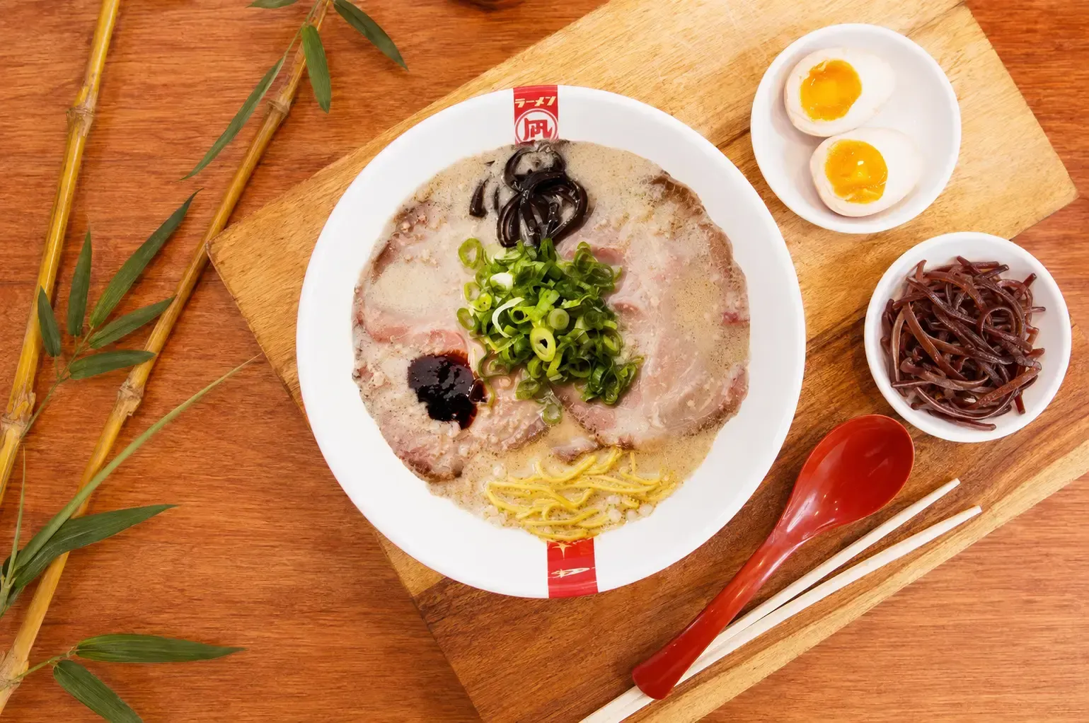 Top-down view of a bowl of Tonkotsu ramen with chashu pork and green onions on a wooden board, served with a soft-boiled egg, wood ear mushrooms, a red spoon, and chopsticks.