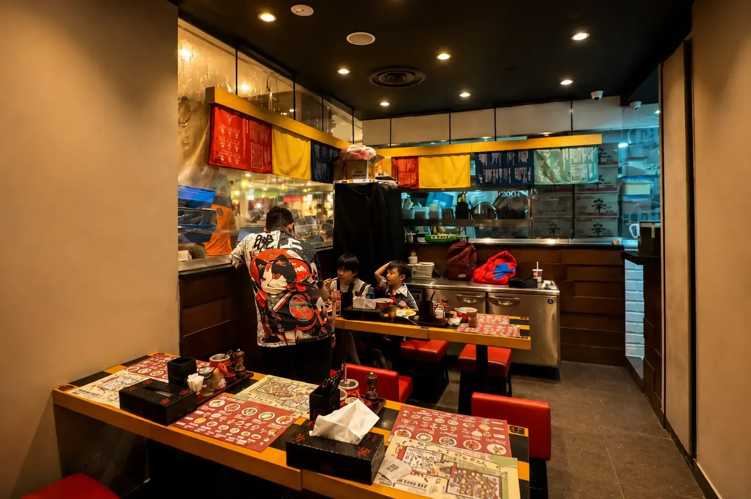 Inside a Ramen Nagi restaurant, showing customers sitting at wooden tables with red stools, menu placemats, and a staff member in a patterned shirt facing the kitchen counter.