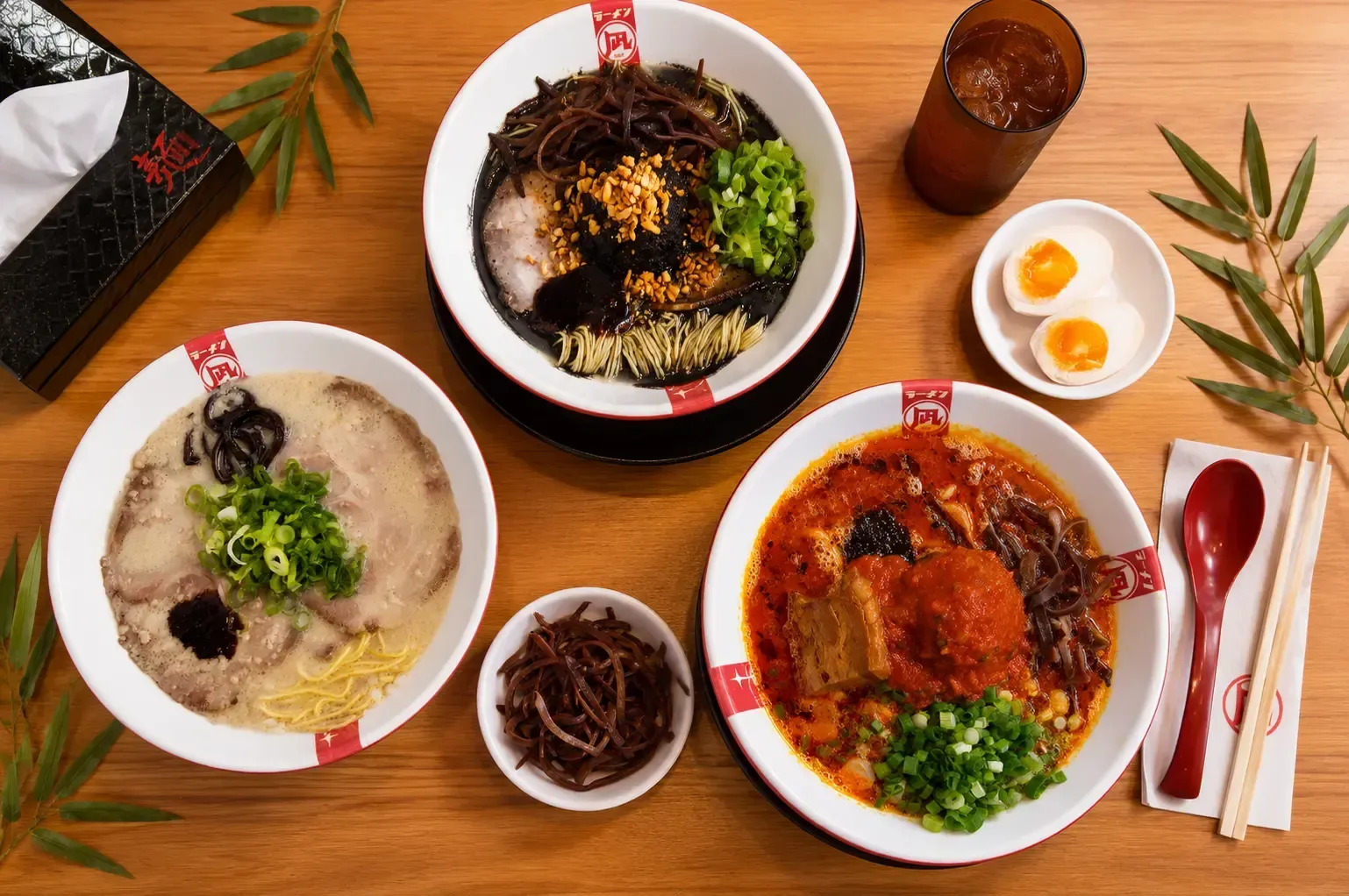 Flat lay view of three different ramen bowls on a wooden table, including a black broth ramen, a white broth ramen, and a red spicy ramen, accompanied by side dishes and drinks.