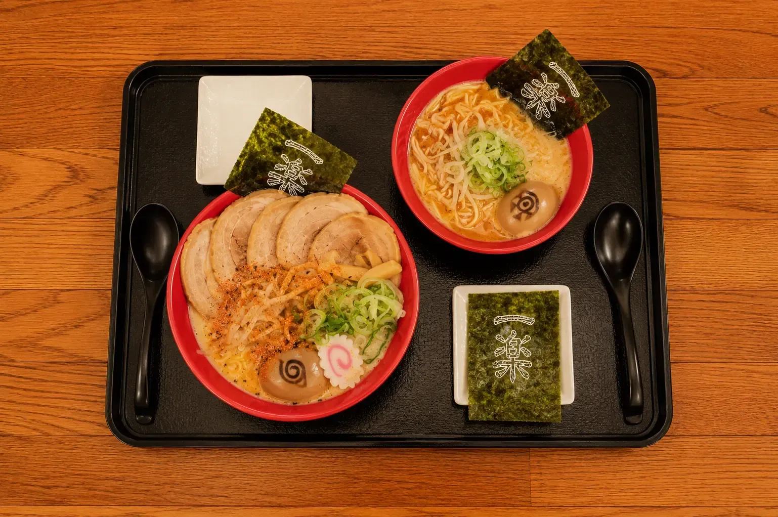 A top-down view of two red bowls of ramen served on a black tray. The larger bowl features several slices of chashu pork, green onions, a fish cake, and a soft-boiled egg with a Naruto spiral symbol, while both bowls include a sheet of nori seaweed branded with white Japanese text.