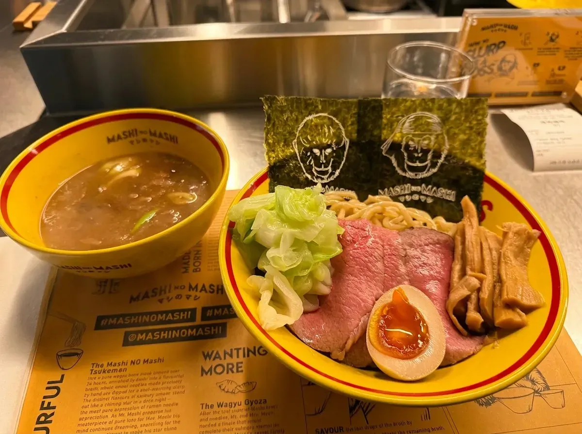 A close-up of a Tsukemen (dipping ramen) dish served in yellow bowls on a paper placemat. The main bowl features thick noodles topped with rare sliced wagyu beef, a soft-boiled egg, cabbage, bamboo shoots, and sheets of nori seaweed printed with illustrated monkey faces. A separate smaller bowl contains the rich dipping broth.