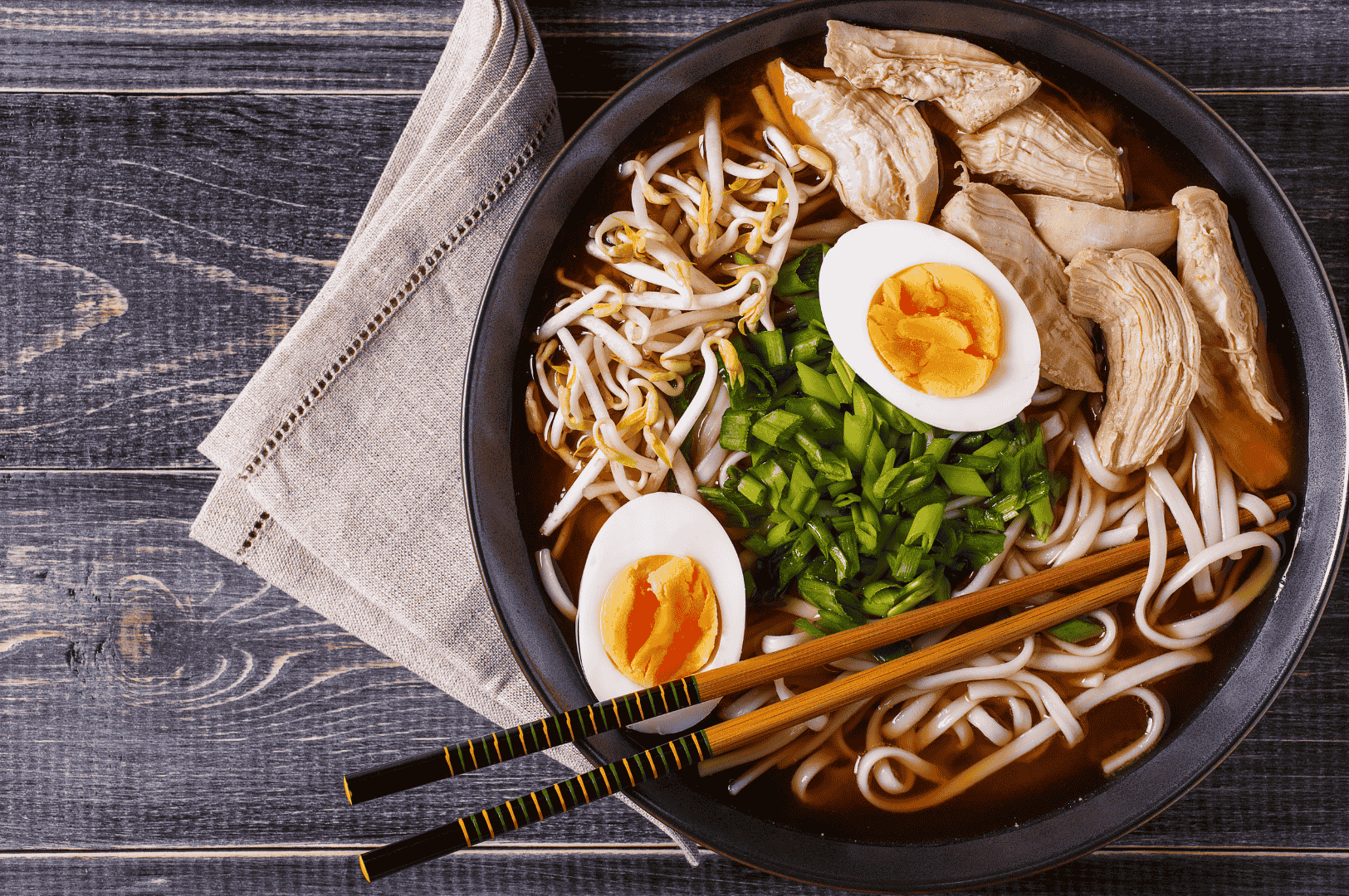 A farm-to-table style ramen bowl in San Francisco, featuring sliced chicken breast, soft-yolk eggs, bean sprouts, scallions, and white noodles in a clear broth. Resting on a dark wooden table with a beige napkin and accented chopsticks, the presentation highlights the artisanal philosophy of Japanese cuisine rooted in freshness, balance, and seasonal ingredients.