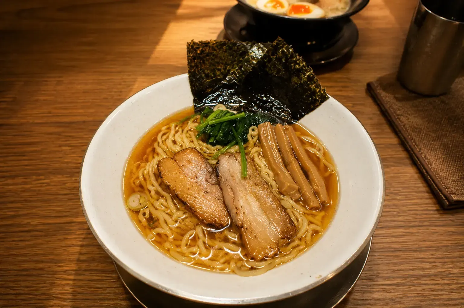 Bowl of soy sauce-based ramen with wavy noodles, thick-cut chashu pork slices, bamboo shoots, greens, and a sheet of nori.